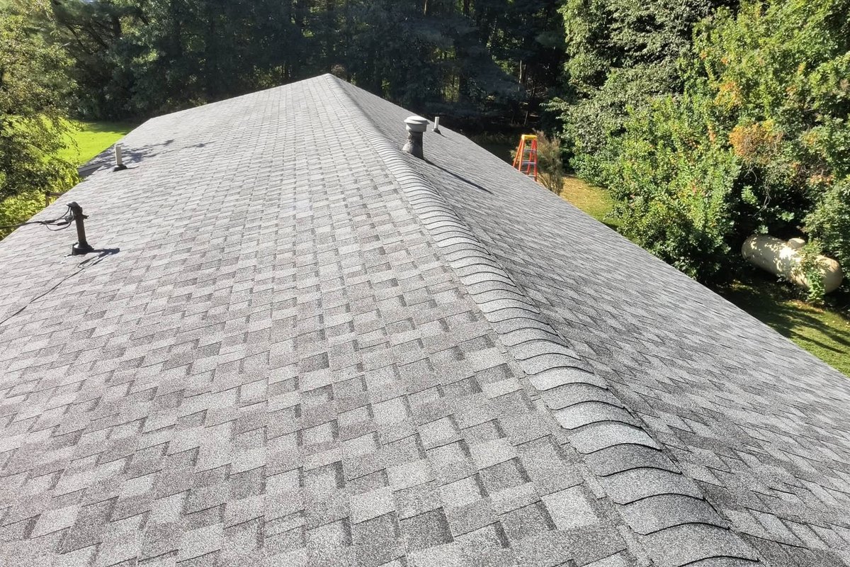 Finished gray asphalt shingle roof with a capped hip, plumbing vents and a stepladder on the lawn beside it